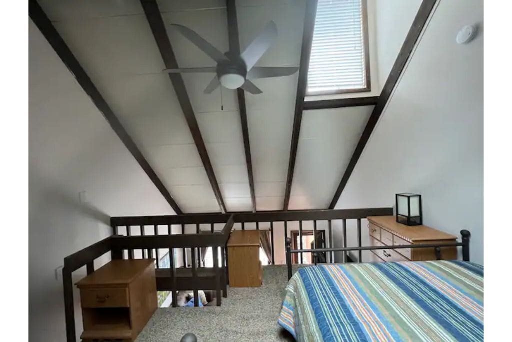 Una stanza con soffitto spiovente, un letto con copriletto a righe, un comodino in legno, una piccola cassettiera in legno e una ventola a soffitto.
