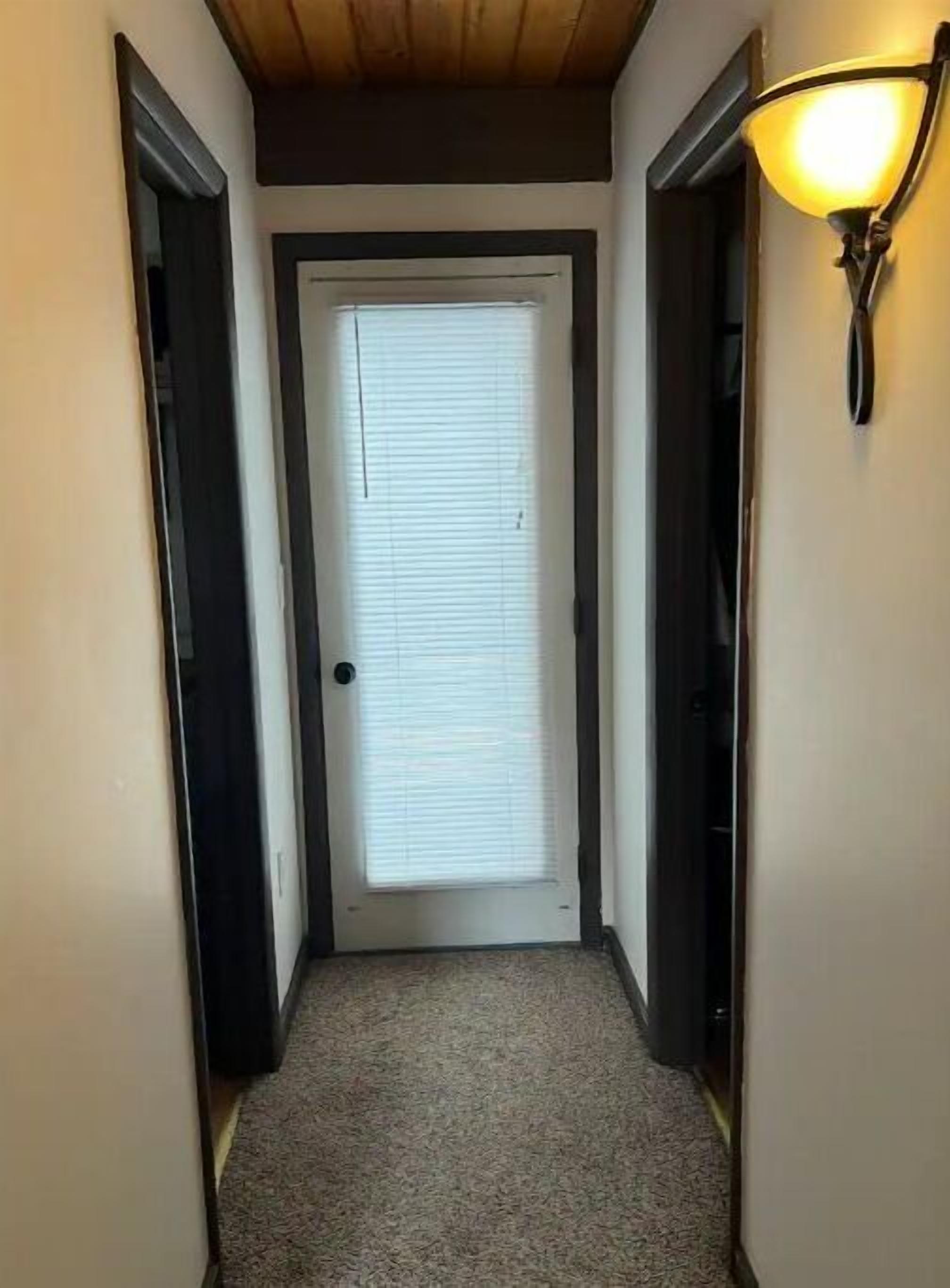 Un couloir étroit, avec une porte au fond, un sol recouvert de moquette et un luminaire fixé au mur.