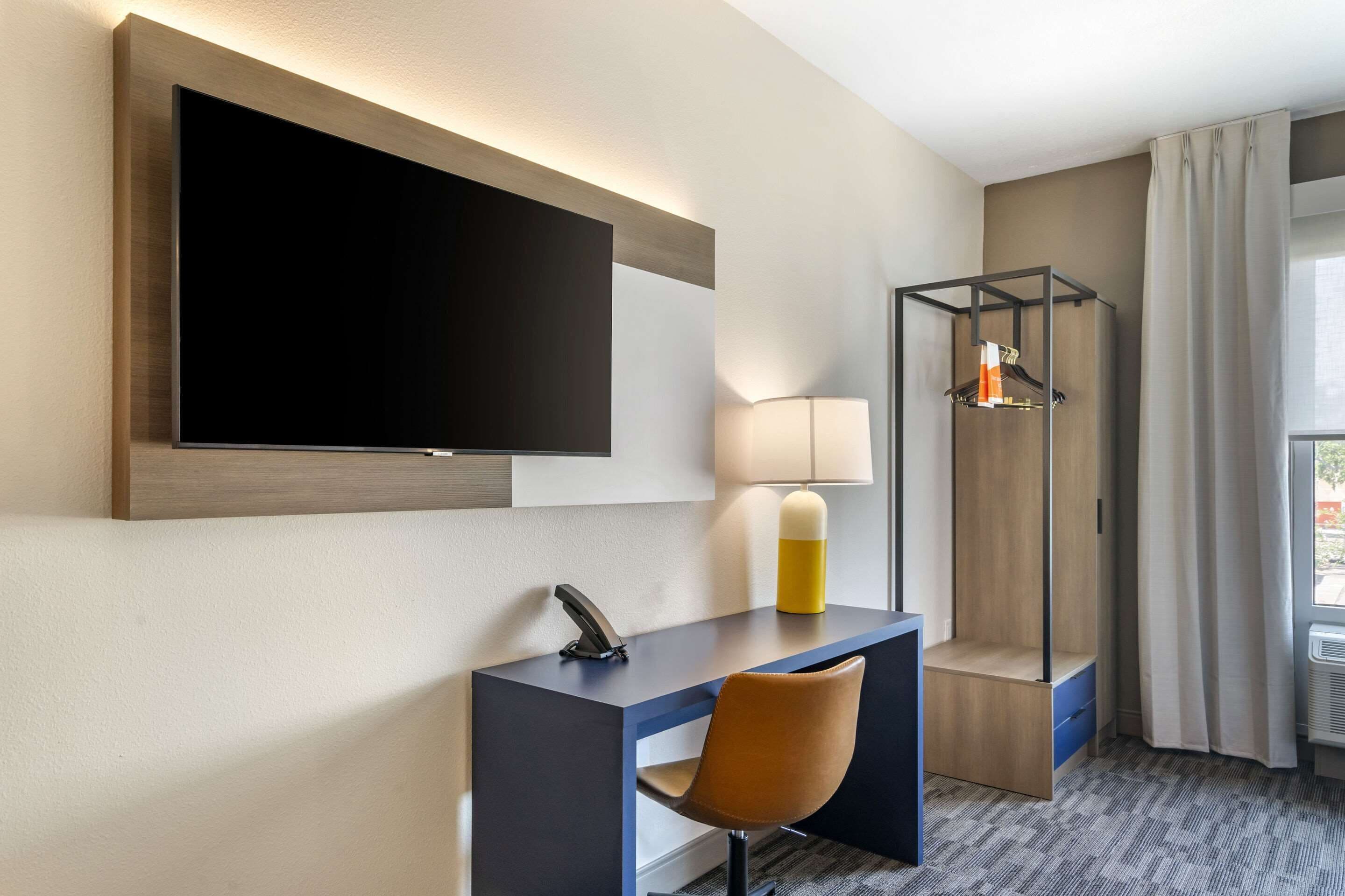 A modern hotel room with a large flat-screen TV mounted on the wall, a desk with a chair, a lamp, and a coat rack.