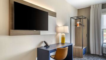 A modern hotel room with a large flat-screen TV mounted on the wall, a desk with a chair, a lamp, and a coat rack.