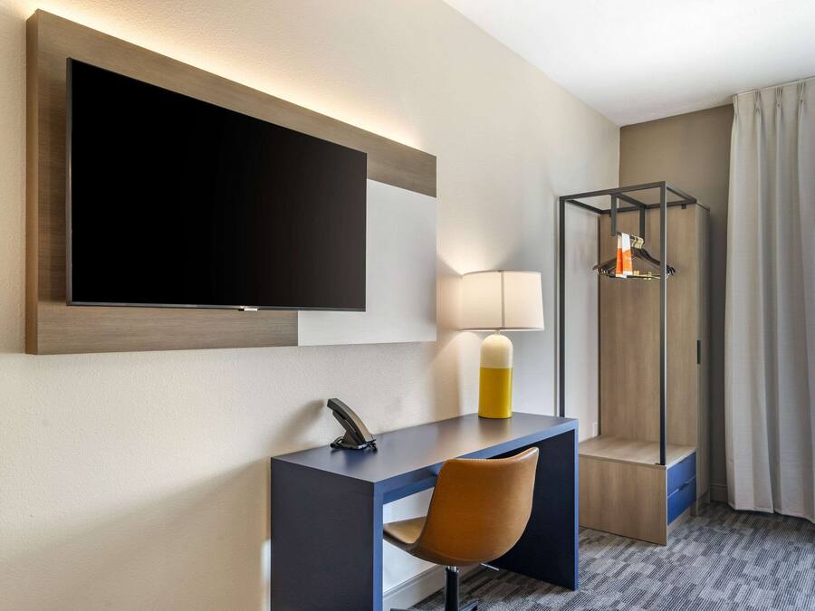 A modern hotel room with a large flat-screen TV mounted on the wall, a desk with a chair, a lamp, and a coat rack.