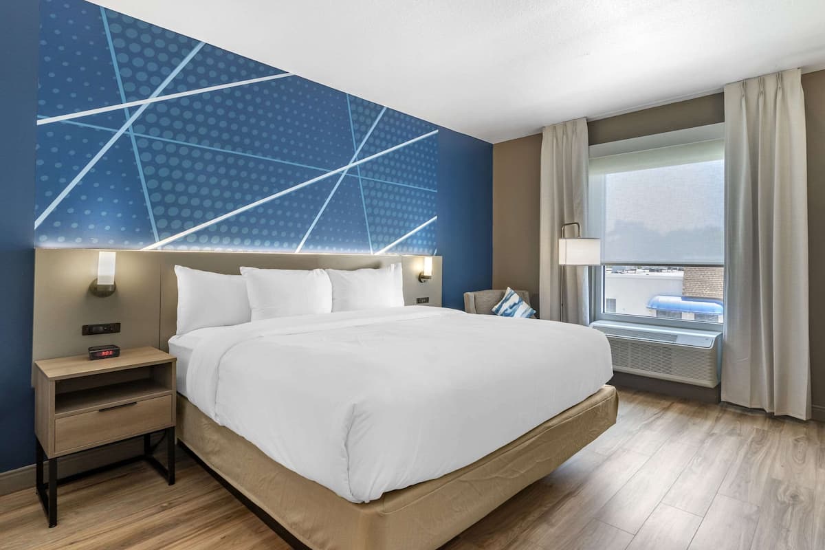 A modern hotel room with a large bed, a nightstand, a wall-mounted lamp, and a window with curtains.