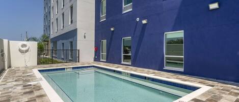 A swimming pool with a tiled deck, a building with a blue facade, and a clear sky.