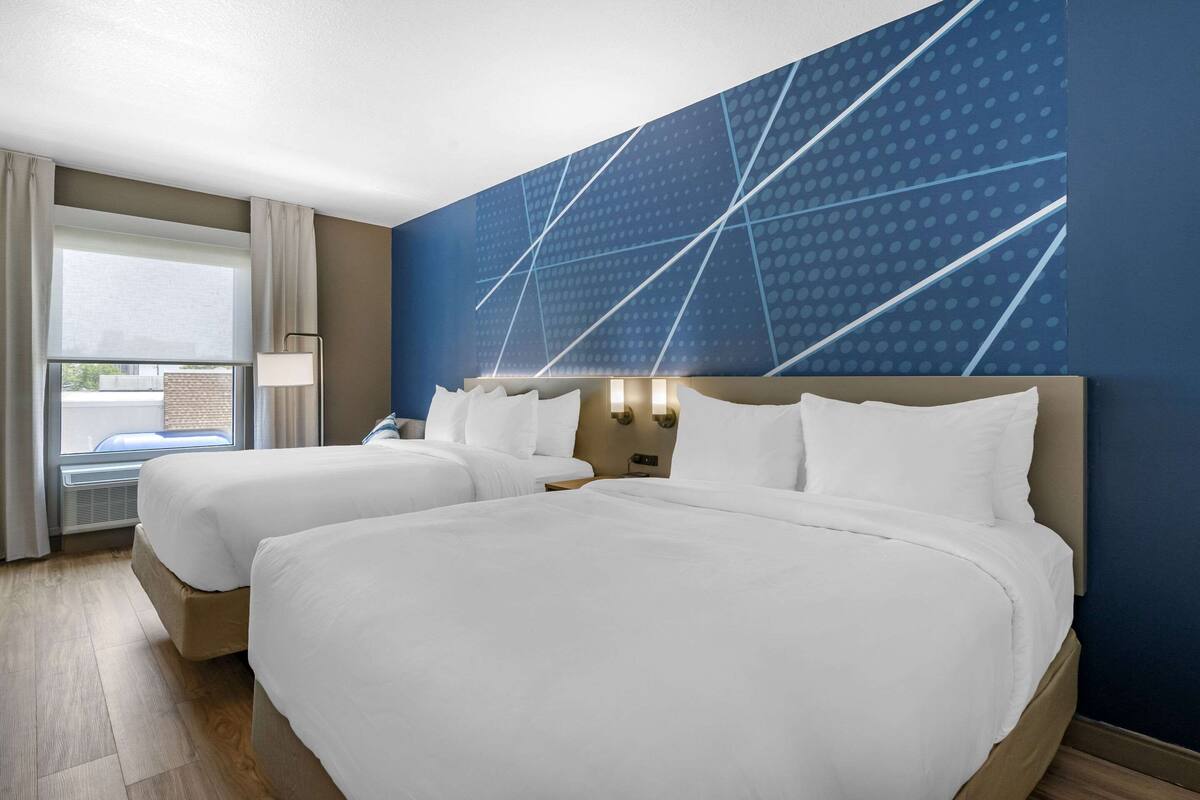 A hotel room with two beds, a large window with curtains, a wall with a geometric pattern, and a nightstand with a lamp.