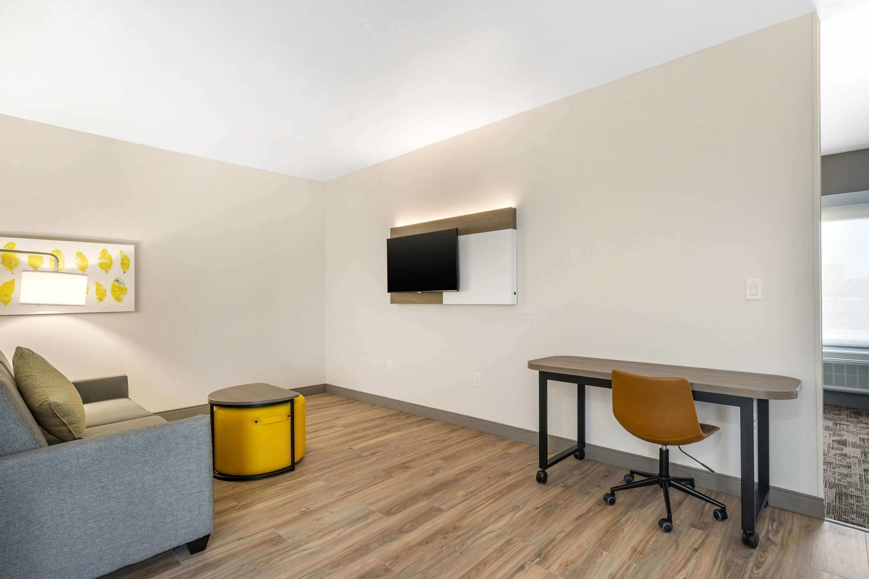 A modern room with a grey sofa, a yellow side table, a wooden floor, a desk with a chair, and a flat-screen TV mounted on the wall.