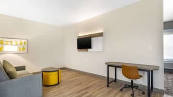 A modern room with a grey sofa, a yellow side table, a wooden floor, a desk with a chair, and a flat-screen TV mounted on the wall.