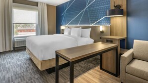 Una habitación de hotel con una cama grande, una mesita de noche de madera, un escritorio y una silla. Hay una ventana con cortinas, una lámpara de pared y un papel tapiz estampado.