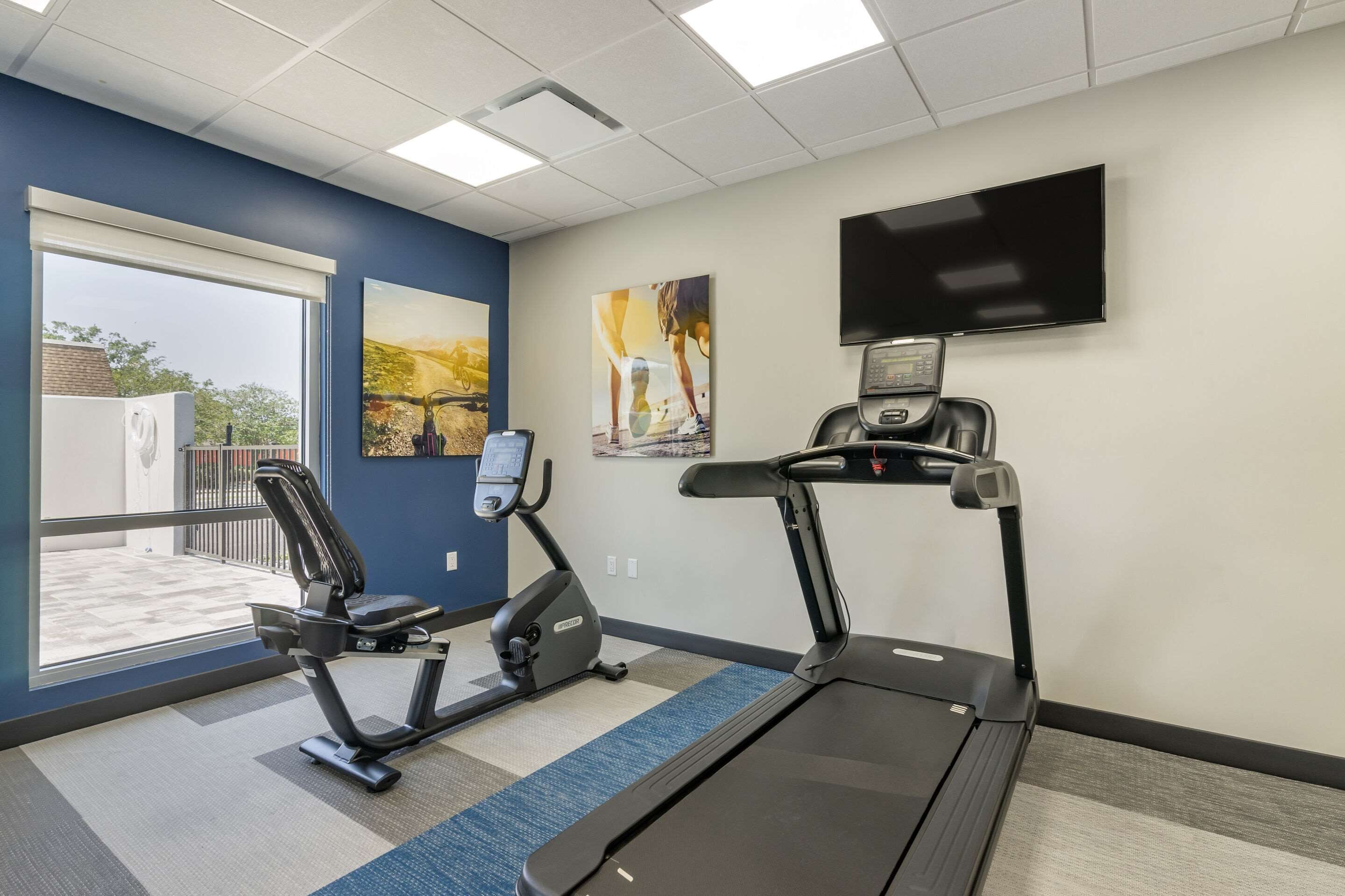 Un gimnasio equipado con cinta de correr, elíptica y bicicleta estática, amplias ventanas y un televisor en la pared.