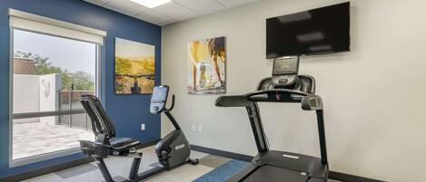 A fitness room with a treadmill, elliptical, and stationary bike, large windows, and a TV on the wall.