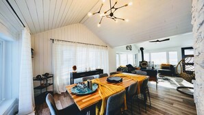 Et hyggeligt, godt oplyst rum med et træspisebord, stole og en moderne lysekrone. En sort komfur ses i baggrunden, og en fletstol er synlig.