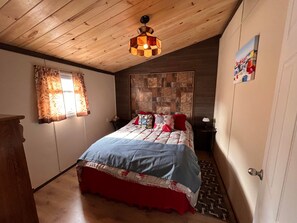 Un dormitorio con techo de madera, colcha acolchada, cabecera estampada, ventana con cortinas y una mesita de noche pequeña.