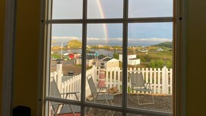 Une vue à travers une fenêtre, avec un arc-en-ciel, des sièges à l’extérieur et une clôture blanche en bois.