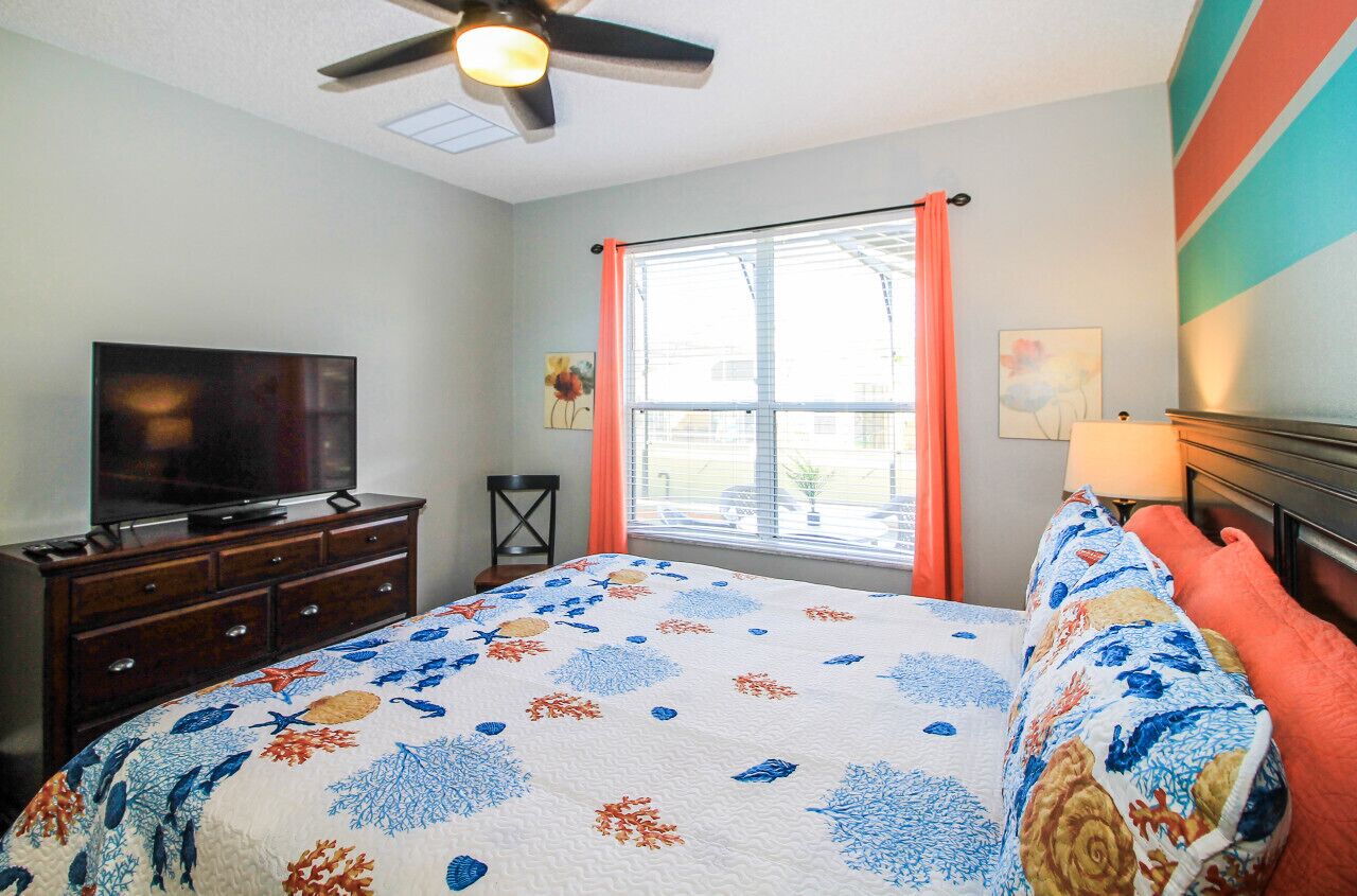 A bedroom with a bed, a TV on a dresser, a window with curtains, and a ceiling fan.