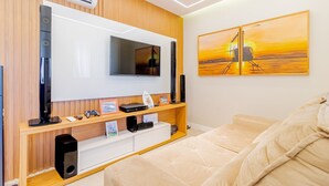 Una habitación de hotel moderna con un televisor de pantalla plana montado en una pared de madera, una mesita consola de madera con dispositivos electrónicos, una cama con ropa de cama color beige y un cuadro de atardecer en la pared.