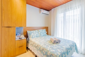 Un dormitorio con un armario de madera, una cama con ropa de cama floral, un equipo de aire acondicionado y cortinas traslúcidas.