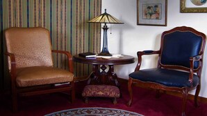 A room with two wooden chairs, one with a cushioned seat and the other with a leather seat, a small round table with a lamp, and a patterned rug on the floor.