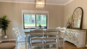 A dining area with a wooden table, white chairs, a chandelier, a window with a view, and a cabinet with drawers.