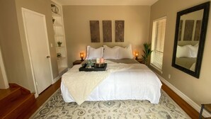 A bedroom with a large bed, two bedside tables with lamps, a mirror, and a door leading to another room.
