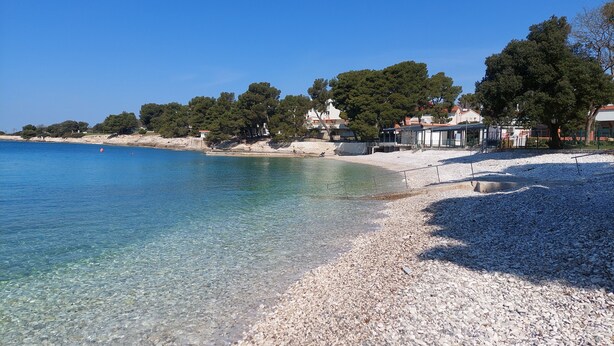 Una playa de guijarros con agua cristalina, algunas personas y edificios al fondo.