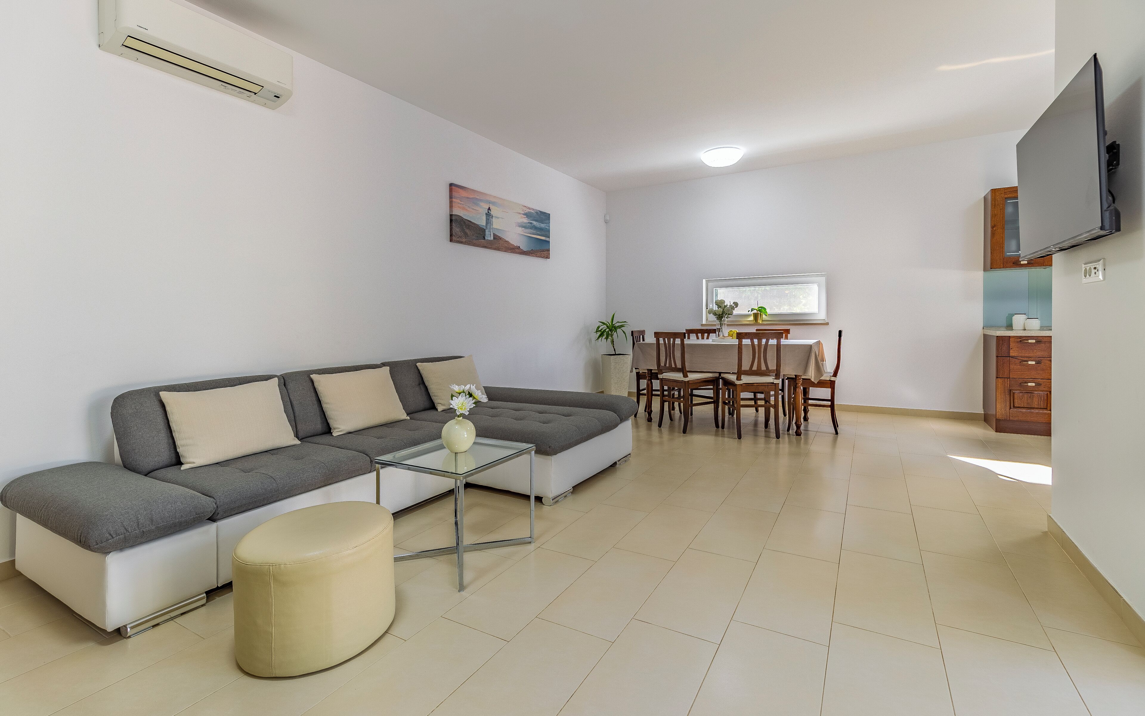 A living room with a grey sofa, a small round ottoman, a glass coffee table, and a painting on the wall.