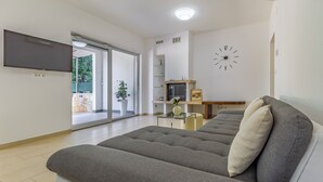 Una sala moderna con un sofá gris, un televisor de pantalla plana montado en la pared y un ventanal con vistas a la vegetación.