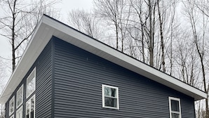 A two-story house with a dark exterior, a stone foundation, and a garage. - Brand new, modern, cozy house in the woods! (Greig)