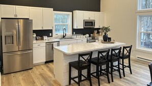 A modern kitchen with white cabinets, stainless steel appliances, a marble countertop, and a large window. - Brand new, modern, cozy house in the woods! (Greig)