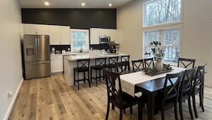 A modern kitchen with white cabinets, a stainless steel refrigerator, and a dining area with a table and chairs. - Brand new, modern, cozy house in the woods! (Greig)