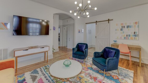 Una sala moderna con un televisor de pantalla plana, una mesa de centro redonda, dos sillones azules, una consola de madera y una alfombra colorida.