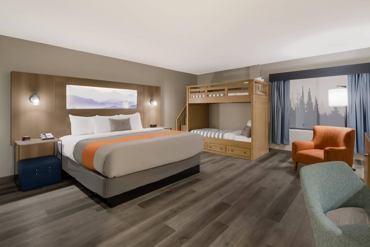 A hotel room with a large bed, a bunk bed, a desk with a lamp, a chair, and a window with curtains.