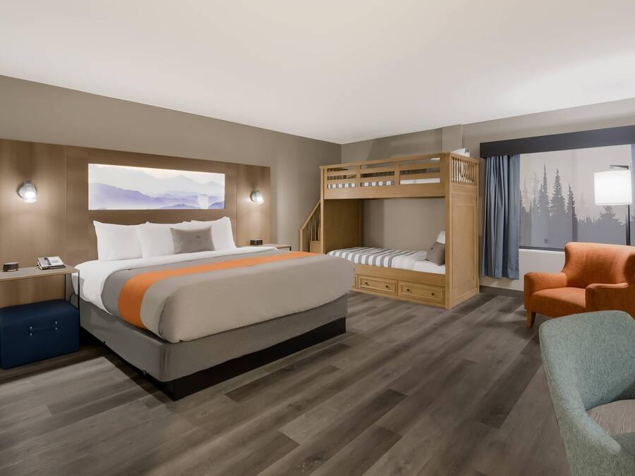 A hotel room with a large bed, a bunk bed, a desk with a lamp, a chair, and a window with curtains.