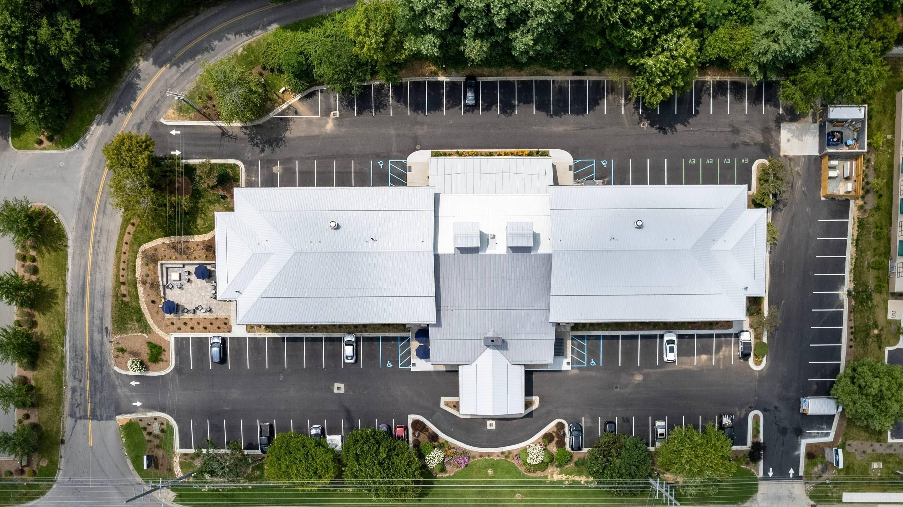 An aerial view of a parking lot with a large building in the center, surrounded by trees and a curved road.