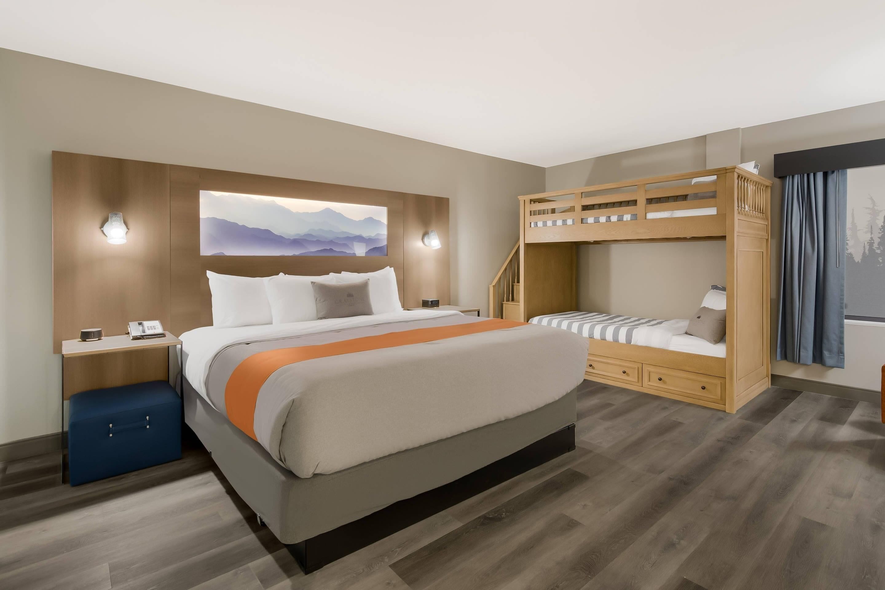Quarto de hotel moderno com uma cama grande, uma beliche, uma escrivaninha com luminária e uma obra de arte na parede.