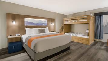 Quarto de hotel moderno com uma cama grande, uma beliche, uma escrivaninha com luminária e uma obra de arte na parede.