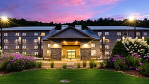 A hotel with a modern architectural design, featuring a central entrance with a covered porch, stone accents, and a well-maintained lawn with flowering plants.