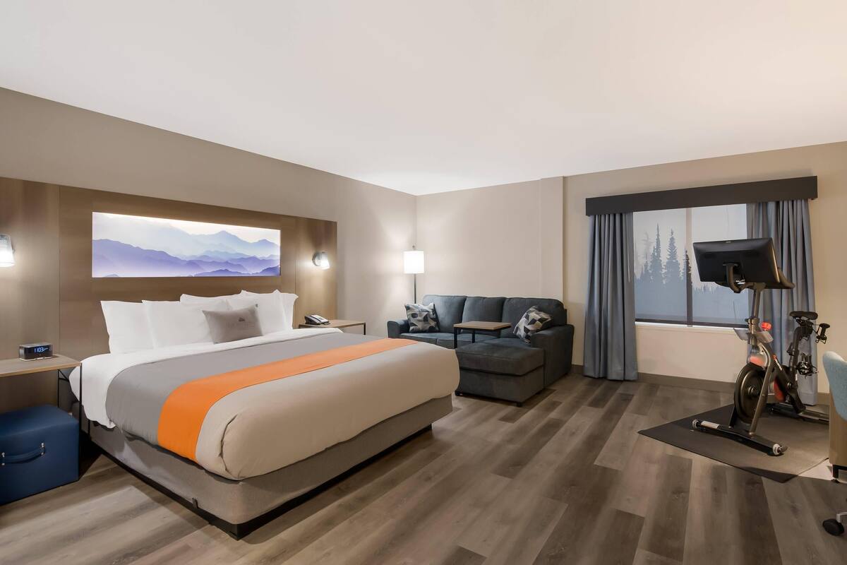 A hotel room with a bed, a sofa, a desk, a lamp, a bike, and a window with curtains.