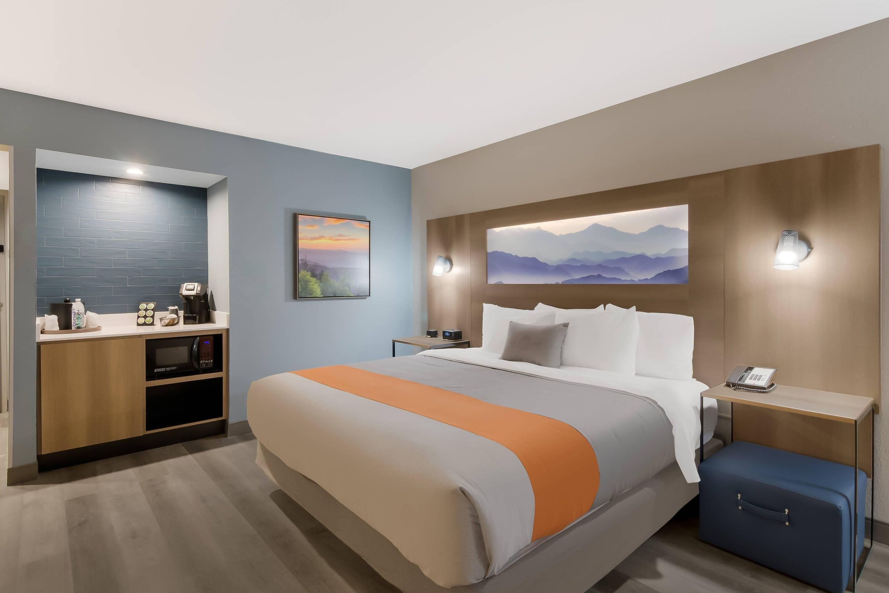 A modern hotel room with a large bed, a desk with a microwave, a phone, and a suitcase.