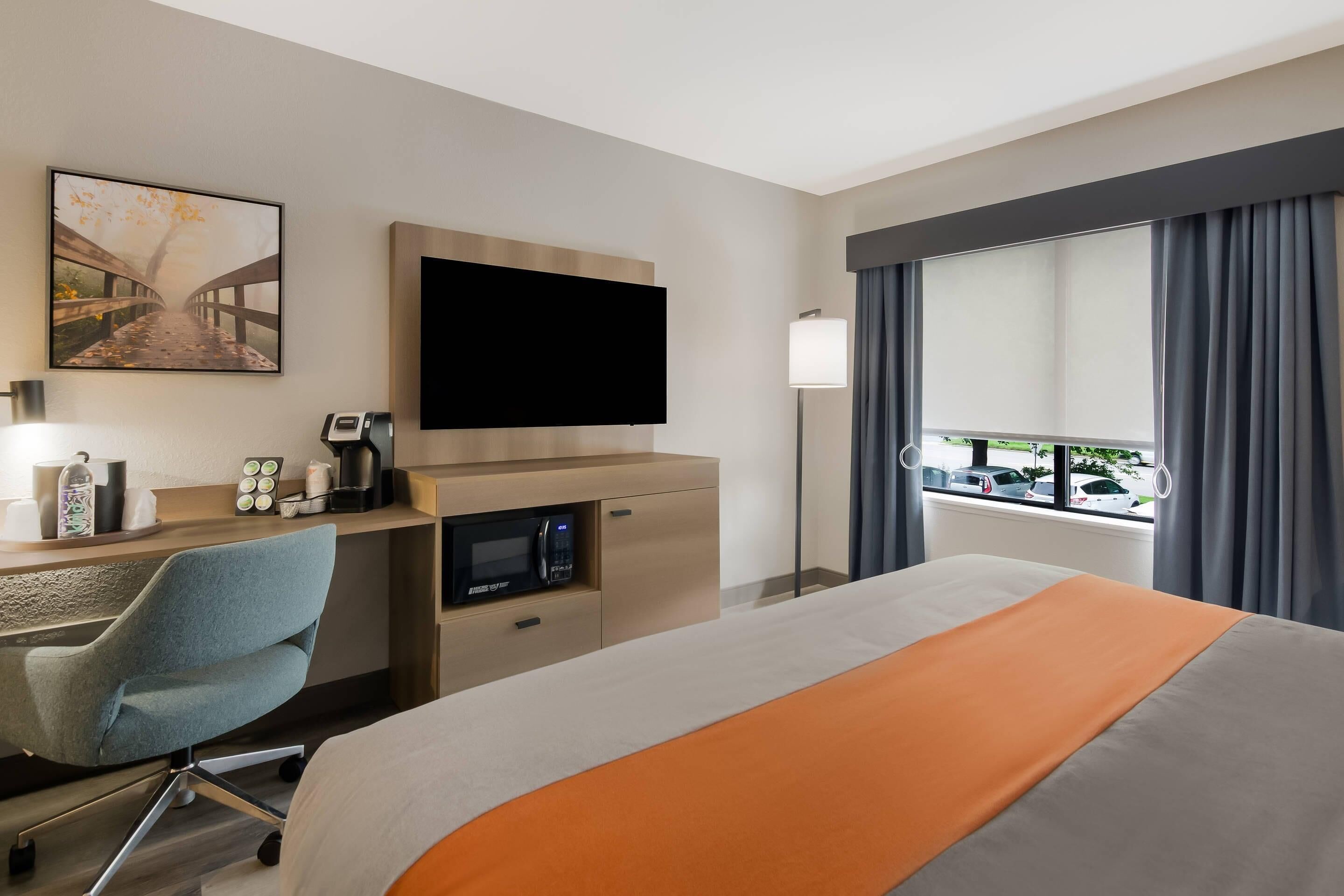 A hotel room with a bed, a desk with a chair, a flat-screen TV, a coffee maker, a microwave, and a window with curtains.