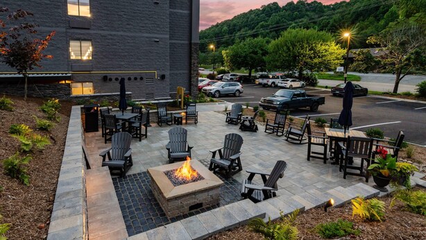 An outdoor patio with fire pit seating and tables, surrounded by landscaped beds and a parking area in the background.