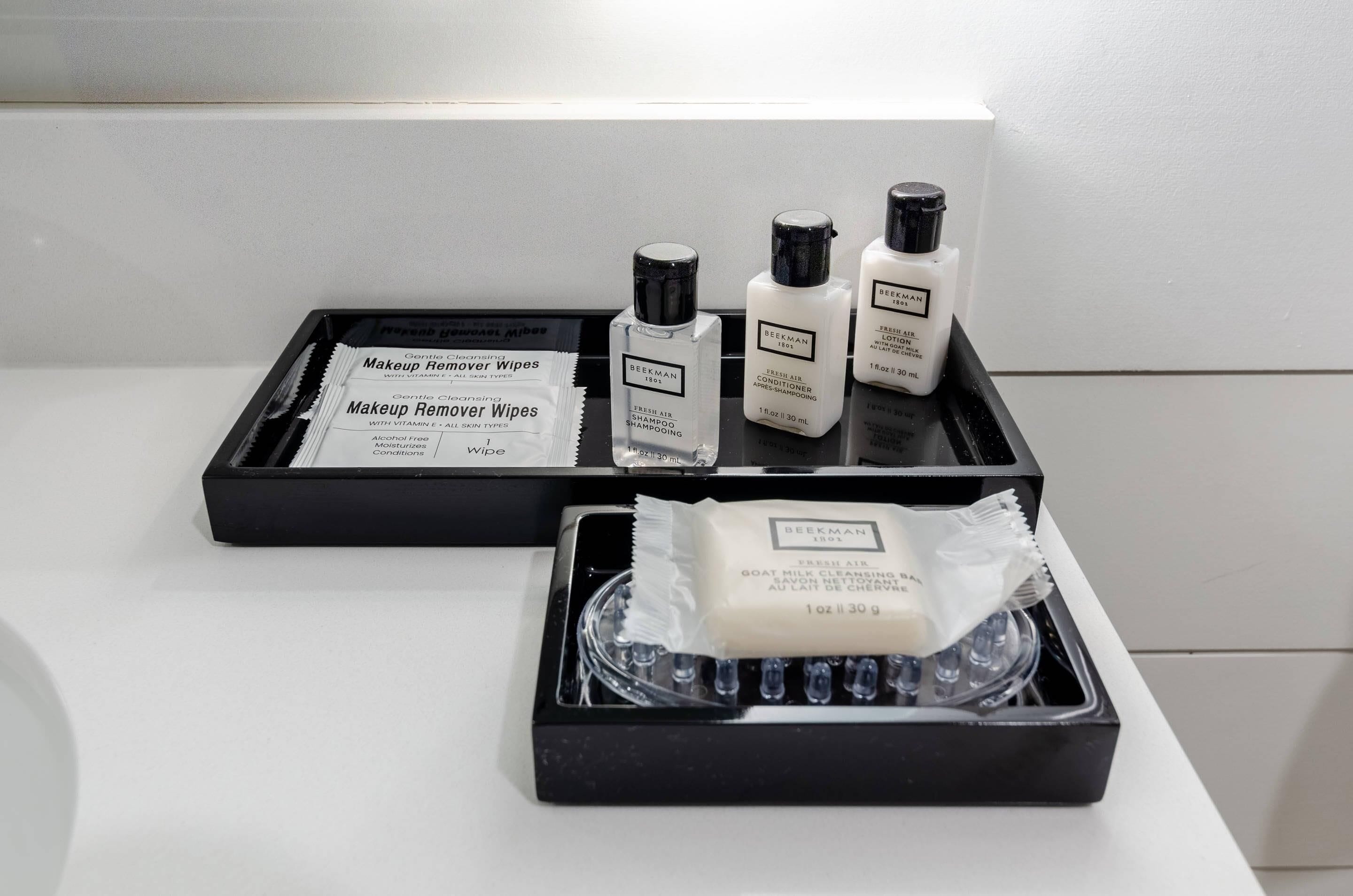 Conjunto de produtos de higiene pessoal, incluindo lenços demaquilantes, condicionador e um sabonete de leite de cabra, tudo em uma bandeja preta.