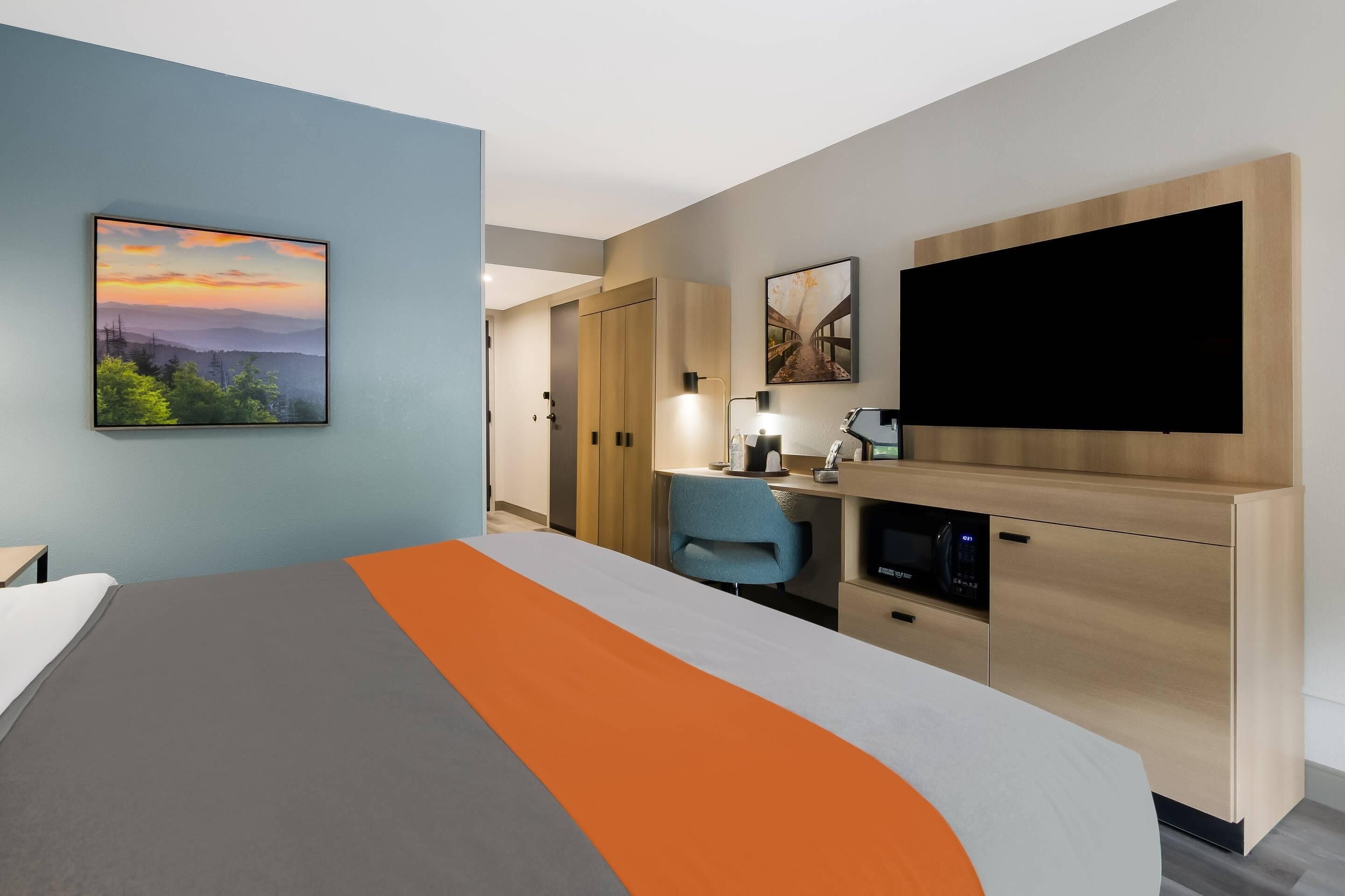 A hotel room with a bed, desk, chair, TV, and artwork on the walls.