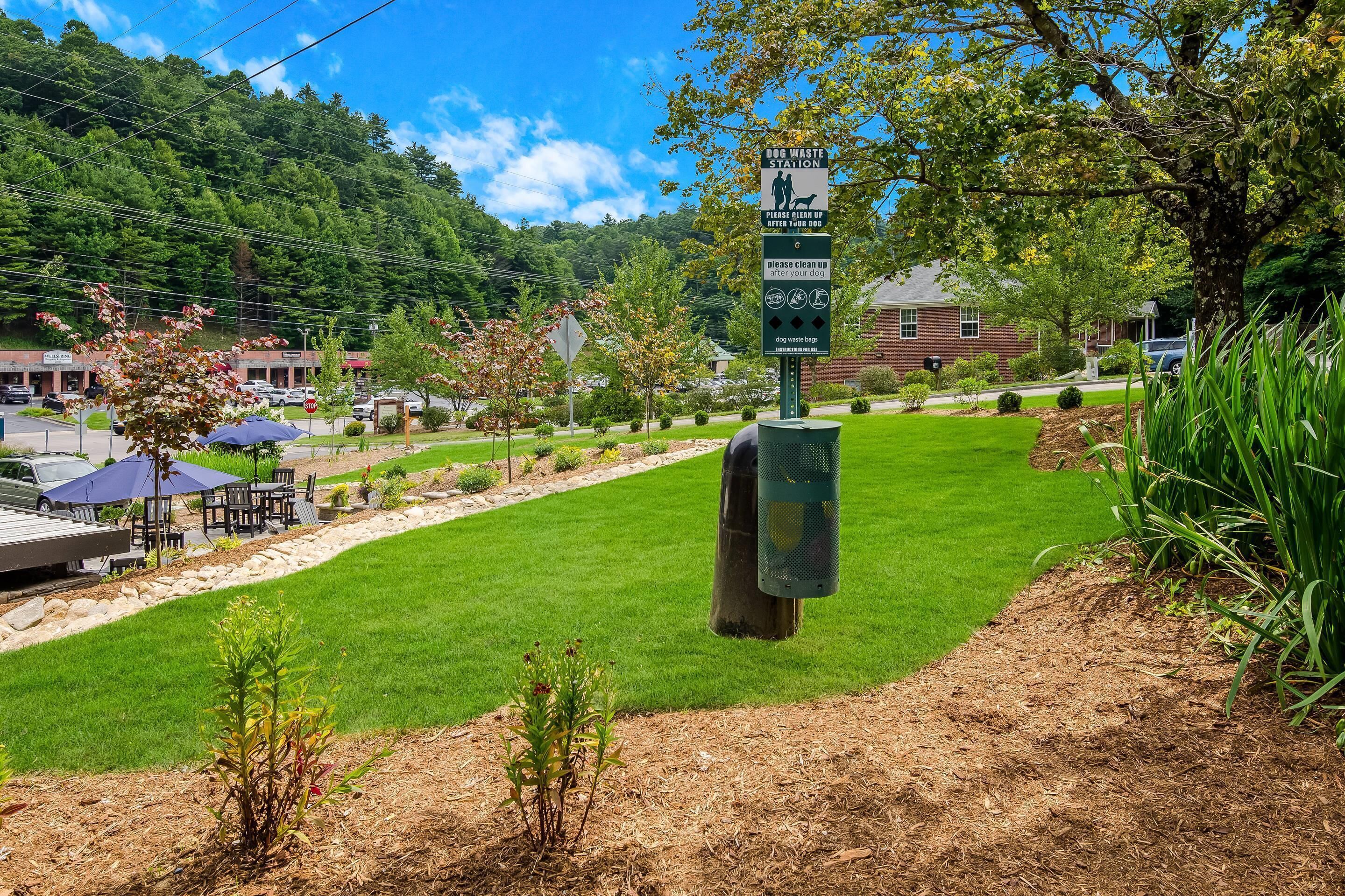 A dog waste station with a sign instructing to clean up after your dog, surrounded by a well-maintained grassy area, trees, and a building in the background.
