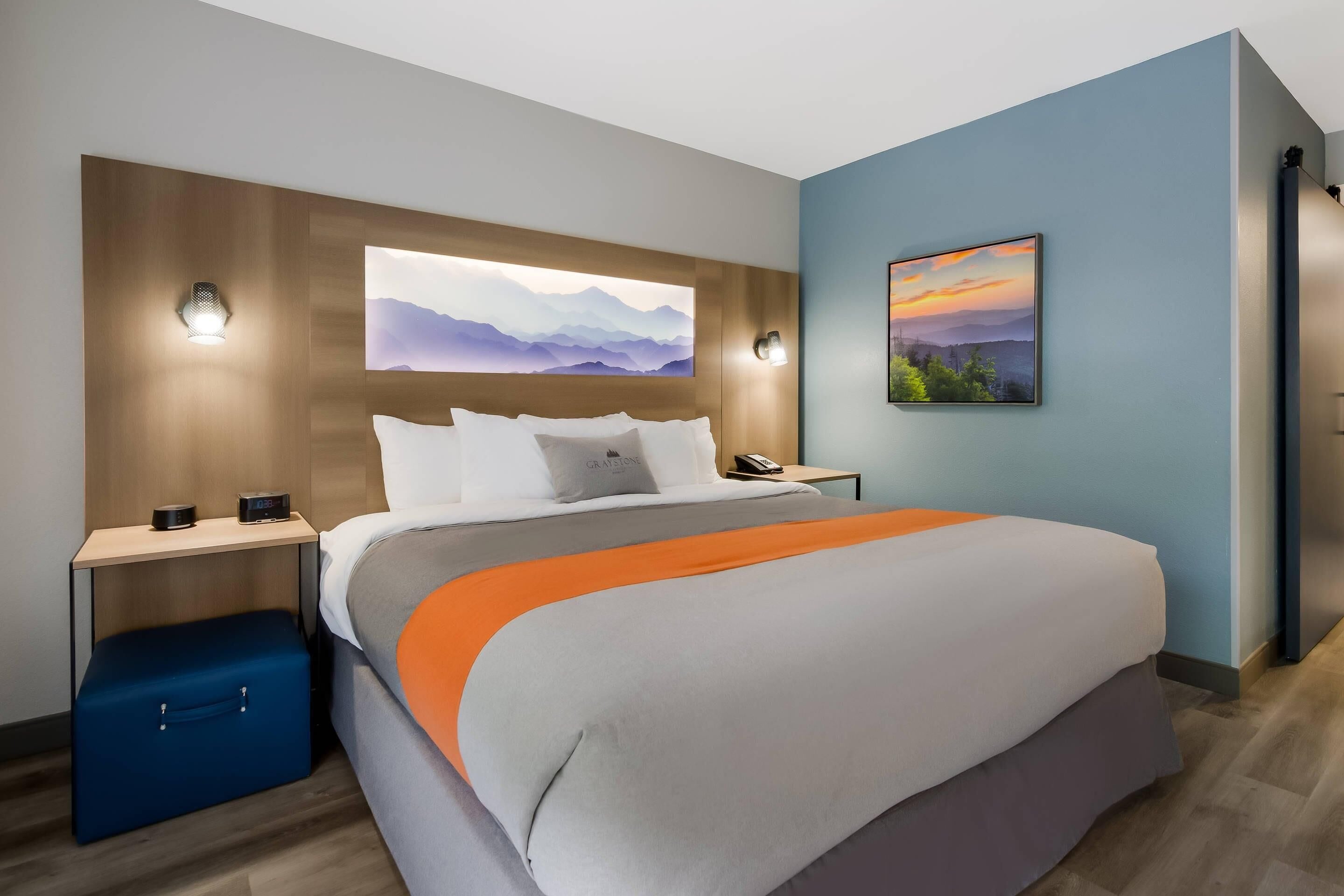 Una habitació d'hotel moderna amb un llit gran, una tauleta de nit amb una làmpada, una maleta blava petita i un quadre penjat a la paret.