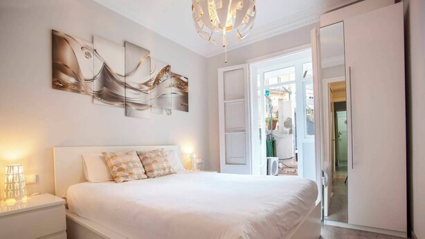 A bedroom with a large bed, a nightstand, a modern chandelier, and a view of the outside through an open door. - P2PAR1002- Super Bright & New 2B Apt in the center (Barcelona)