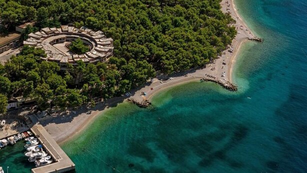 Luftaufnahme einer Küstenlandschaft mit einem kreisförmigen Gebäude, umgeben von dichtem Baumbestand, einem Sandstrand und klarem, blauem Wasser.