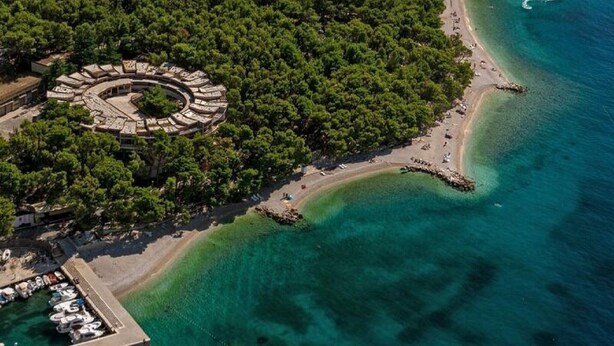 Luftaufnahme einer Küstenregion mit einer kreisförmigen Struktur, umgeben von dichtem Baumbestand, einem Strand und klarem, blauem Wasser.