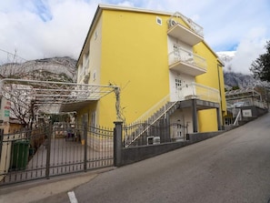 A yellow building with a staircase leading to a balcony, surrounded by a metal fence and a paved area. - Apartments Pezo - Studio with Balcony and Sea View (Baška Voda)