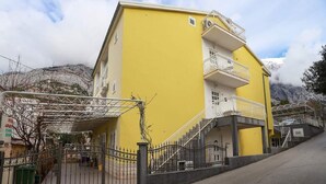 A yellow building with a staircase leading to a balcony, surrounded by a metal fence and a paved area. - Apartments Pezo - Superior Two Bedroom Apartment with Balcony (Baška Voda)