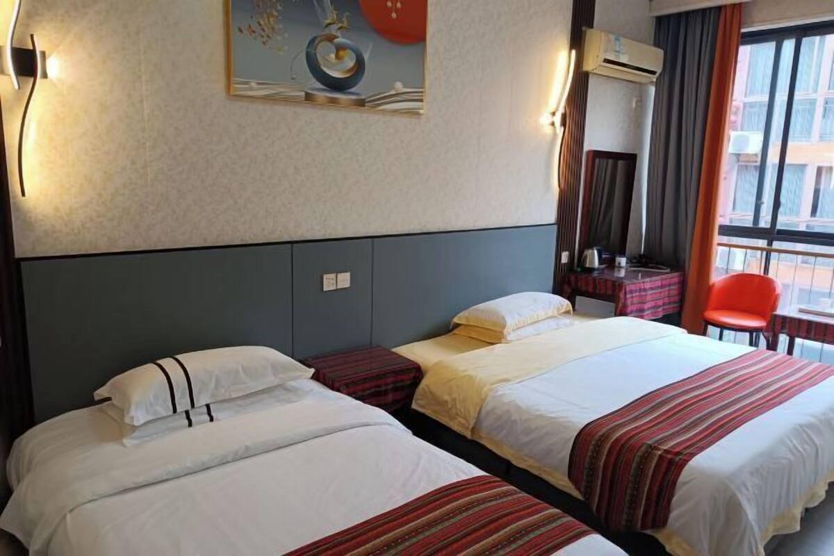 Habitación de hotel con dos camas, una mesita, una silla y un ventanal con cortinas.