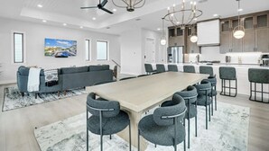 Een moderne woonkamer met een grote houten eettafel, grijs gestoffeerde stoelen, een grijze bank en een kroonluchter.